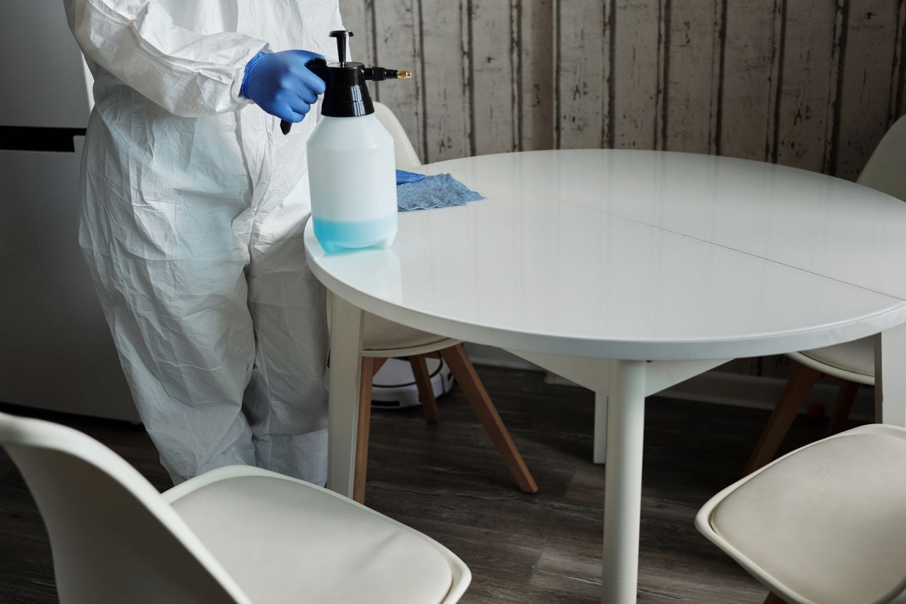 Personne en combinaison de protection désinfectant une table blanche à l'intérieur avec un pulvérisateur.